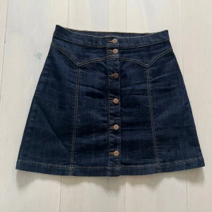 Mörkblå jeanskjol med knappar - Snygg mörkblå jeanskjol från H&M med knappar framtill. Kjolen har en klassisk A-linjeform och dekorativa sömmar som ger en stilren look. Jeanskjol med knäppning fram. St. 38 Längd 44 cm Midja 2 x 37-38 cm H&M Ny, endast tagg borttagen och provad.