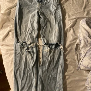 Helutsvängda baggy jeans med slitningar - Säljer ett par ljusblå jeans med slitningar över knäna. De har en klassisk femficksdesign och en bekväm passform. Perfekta för en avslappnad stil. Helutsvängda vilket är välldigt trendigt nu och i ny skick