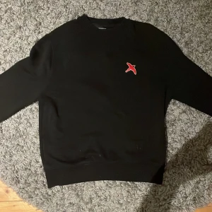 Svart tröja från Axel Arigato - Säljer en stilren svart tröja från Axel Arigato med en broderad röd fågel på bröstet. Tröjan har en klassisk passform och är perfekt för en avslappnad look. Den är tillverkad i ett mjukt material som ger en skön känsla. Nypris 2000