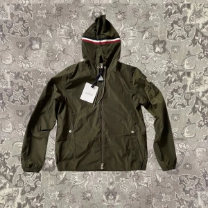 Grön vindjacka från Moncler - Snygg grön vindjacka från Moncler med huva och dragkedja. Jackan har en röd och vit rand på huvan och elastiska ärmslut. Perfekt för blåsiga dagar.