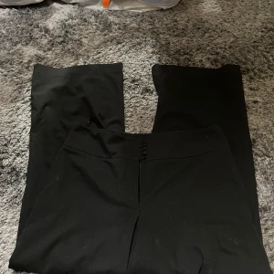 Svarta kostymbyxor från Hennes - Snygga svarta kostymbyxor från Hennes med en elegant design. Byxorna har en rak passform och en diskret knappdetalj baktill. Perfekta för en stilren look.