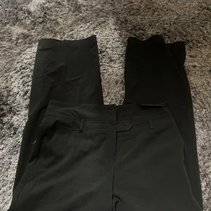 Svarta kostymbyxor från KappAhl - Snygga svarta kostymbyxor från KappAhl med en klassisk design. Byxorna har en rak passform och är perfekta för en stilren look. De har en diskret dragkedja och knapp framtill.