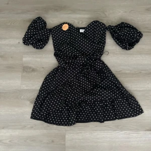 Svart prickig klänning - Charmig svart klänning med vita prickar och puffiga kortärmar. Klänningen har en volangdetalj vid fållen och ett knytband i midjan för en smickrande passform. Perfekt för en stilfull look.