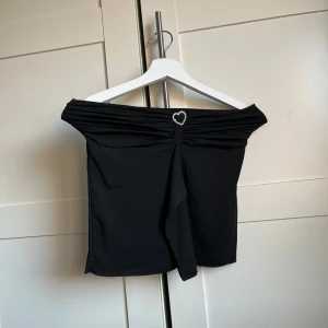Svart offshoulder topp med hjärtdetalj - Snygg svart offshoulder topp med en glittrig hjärtdetalj framtill. Perfekt för en kväll ute eller en speciell tillställning. Toppen har en elegant drapering och är tillverkad i ett mjukt material.