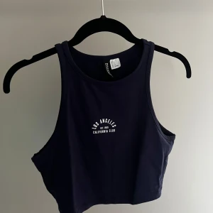Mörkblå croppad linnetopp från Divided - Säljer en mörkblå croppad linnetopp från Divided med tryck 'Los Angeles California Club' på framsidan. Perfekt för en avslappnad stil. Toppen är ärmlös och har en rund halsringning.