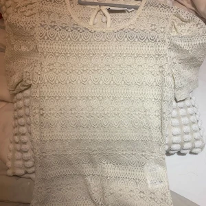 Vit spetsblus - Säljer en elegant vit spetsblus med korta ärmar och rund hals. Blusen har ett vackert spetsmönster och en knytning i nacken. Perfekt för en stilren look.