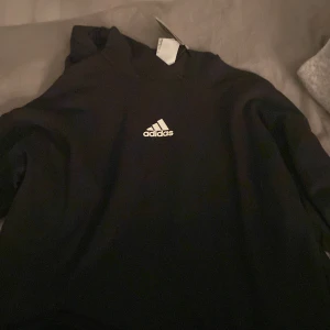 Svart hoodie från Adidas - Säljer en klassisk svart hoodie från Adidas med deras ikoniska logga på bröstet. Perfekt för en avslappnad stil. Den har långa ärmar och en bekväm passform.