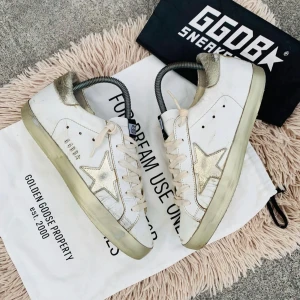 Golden Goose sneakers med guldstjärna - Snygga Golden Goose sneakers i vitt med en ikonisk guldstjärna på sidan. Skorna har en vintage-look med snörning och gulddetaljer på hälen. Perfekta för en trendig och avslappnad stil.