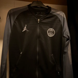 Svart jacka från Nike x Jordan - Snygg svart jacka från Nike i samarbete med Jordan och PSG. Jackan har Dri-FIT-teknologi och dragkedja framtill. Den har långa ärmar och ett stort 'Paris' tryck på ryggen. Perfekt för sportiga tillfällen.