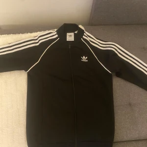 Svart träningsjacka från Adidas - Säljer en klassisk svart träningsjacka från Adidas med vita ränder längs ärmarna och dragkedja framtill. Jackan har en normal passform och är perfekt för sportiga aktiviteter. Den ikoniska Adidas-loggan finns på bröstet.