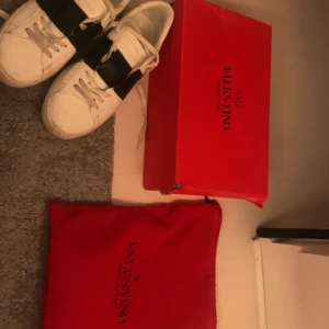 Valentino sneakers med svart detalj - Snygga vita Valentino sneakers med en bred svart rem över snörningen. Skorna har en klassisk design med rund tå och är tillverkade i skinn. Perfekta för en stilren look.