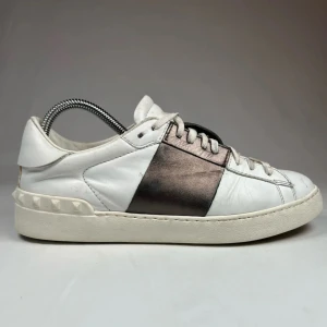 Valentino sneakers i vitt och svart - Snygga Valentino sneakers i vitt skinn med en bred glittrig rem över ovansidan. Skorna har klassisk snörning och en bekväm sula. Perfekta för en stilren look.