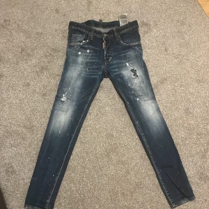Blå jeansbyxor från DSQUARED2 - Snygga blå jeansbyxor från DSQUARED2 med slitna detaljer och färgstänk. De har en slim passform och klassisk femficksdesign. Perfekta för en trendig look.