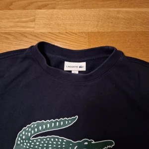 Mörkblå tröja från Lacoste - Säljer en snygg mörkblå tröja från Lacoste med det ikoniska krokodilmärket på framsidan. Tröjan har långa ärmar och är perfekt för en stilren look. Passar bra till både jeans och chinos.