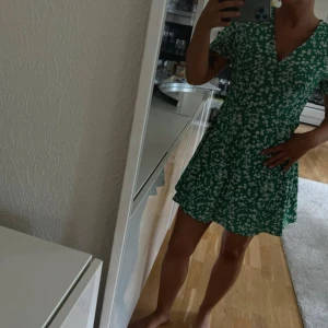 Grön blommig klänning - Säljer en söt grön klänning med vita blommor. Klänningen har en v-ringad design och korta ärmar, perfekt för sommarens varma dagar. Den är kort och har en avslappnad passform som ger en skön känsla. Perfekt för en dag ute i solen!