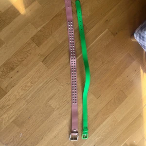 Två bälten i grönt och rosa - Säljer två bälten, ett grönt och ett rosa. Det gröna bältet har en enkel design med en plastspänne och märket TRN 1961. Det rosa bältet har silverfärgade nitar längs hela längden och ett metallspänne. Perfekt för att ge din outfit en färgklick! 50kr styck