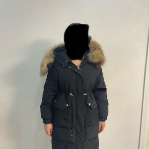 Svart parkajacka med pälsdetaljer - Snygg svart parkajacka med stor huva och pälsdetaljer. Jackan har dragkedja och knappar framtill samt justerbar midja för perfekt passform. Perfekt för kalla dagar.