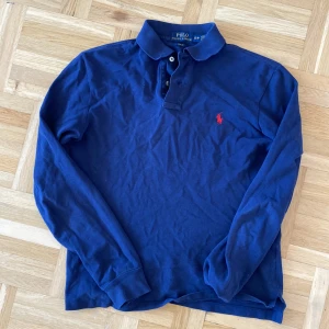 Blå långärmad pikétröja från Ralph Lauren - Säljer en stilren blå långärmad pikétröja från Ralph Lauren. Tröjan har en klassisk krage och knappar framtill. Den är slim fit och har det ikoniska röda broderade logotypen på bröstet. Perfekt för en avslappnad men ändå elegant look. 