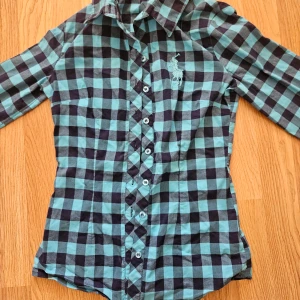 Rutig skjorta från Ralph Lauren - Snygg rutig skjorta i blått och svart från Ralph Lauren. Skjortan har långa ärmar och knappar framtill. Den är dekorerad med det klassiska broderade logotypen på bröstet. Perfekt för en stilren look! Materialet är lite stretchig