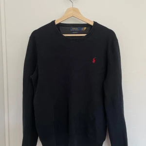 Svart tröja från Ralph Lauren - Säljer en stilren svart tröja från Ralph Lauren i mjuk pima bomull. Tröjan har en klassisk design med rund hals och det ikoniska röda logotypbroderiet på bröstet. Perfekt för en avslappnad men snygg look.