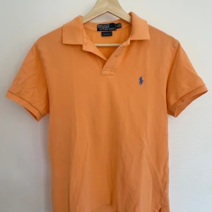 Orange pikétröja från Ralph Lauren - Säljer en snygg orange pikétröja från Ralph Lauren i slim fit. Tröjan har korta ärmar och en klassisk krage med knappar. Den ikoniska loggan är broderad på bröstet i blått. Perfekt för en stilren och avslappnad look. Den är lite mer aprikos orange än vad som syns på bilden. Sitter mer som S men funkar som M