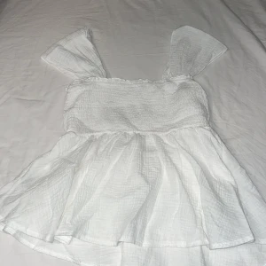 Vit ärmlös topp - Säljer en söt vit topp med smockdetaljer och volangkant. Toppen är ärmlös med breda axelband och har en luftig passform nedtill. Perfekt för varma dagar!