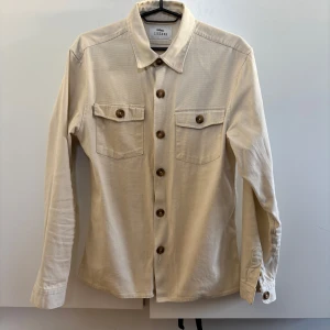 Beige overshirt från Ciszere - Snygg beige overshirt från Ciszere med långa ärmar och knappar framtill. Skjortan har två bröstfickor med knappar och en klassisk krage. Perfekt för lager-på-lager-stil. Stl. L men passar M