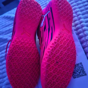 Rosa fotbollsskor från Puma - Säljer ett par rosa fotbollsskor från Puma med svarta ränder och snörning. Skorna har en mönstrad sula för bra grepp på planen. Perfekta för den som vill sticka ut med stil på fotbollsplanen.