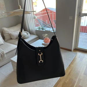 Svart axelväska med gulddetalj - Snygg svart axelväska i skinn med en elegant gulddetalj på framsidan. Väskan har en praktisk dragkedja upptill och en justerbar axelrem. Perfekt för att bära dina dagliga nödvändigheter med stil. Köpte den i London men har aldrig använt den, nypris 399