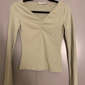 Grön ribbad topp från NA-KD - Säljer en stilren grön ribbad topp från NA-KD med v-ringning och rynkdetalj framtill. Toppen har långa ärmar och en figurnära passform. Perfekt för en trendig look!