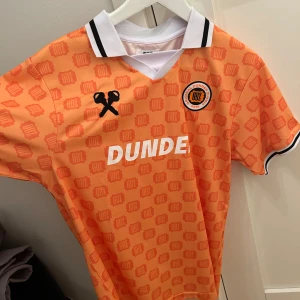 Orange fotbollströja från ODZ - Snygg orange fotbollströja från ODZ med ett unikt mönster och vit text. Tröjan har korta ärmar och en vit krage med svarta detaljer. Perfekt för fotbollsfans som vill sticka ut på planen! Limiterad modell svår att få tag i