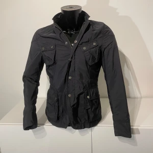 Mörkblå J.lindberg jacka - Snygg mörkblå jacka från J.lindberg med flera fickor och knappar. Jackan har en stilren design med långärmade ärmar och en hög krage. Perfekt för en cool och elegant look.