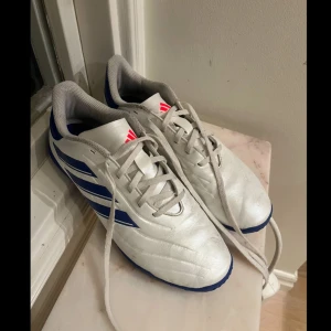 Vita fotbollsskor från Adidas - Säljer ett par vita fotbollsskor från Adidas med blå detaljer och snörning. Skorna har en klassisk design med tre blå ränder på sidan och en neon rosa logotyp på plösen. Fotbollsskorna är endast använd 1 halv säsong (två månader) och används inomhus (futsal).