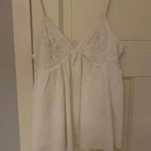 Vit spetslinne - Säljer ett sött vitt linne med spetsdetaljer på bysten och tunna axelband. Perfekt för varma sommardagar eller som ett lager under en kofta. Linnet har en luftig passform och är tillverkat i ett mjukt material.