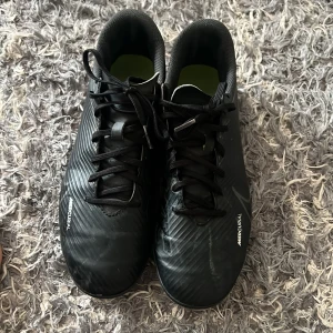 Svarta fotbollsskor från Nike - Säljer ett par svarta Nike Mercurial fotbollsskor med snörning. Skorna har en stilren design med diskreta detaljer och är perfekta för fotbollsplanen. De har en låg profil och är gjorda för att ge bra grepp och kontroll. Har använts bara ett par månader 