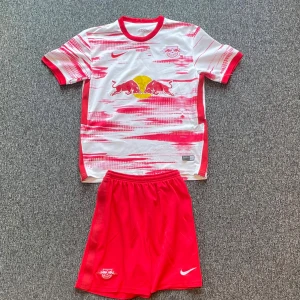 RB Leipzig fotbollströja och shorts - Säljer en RB Leipzig fotbollströja med matchande shorts. Tröjan är vit med röda detaljer och har korta ärmar. Den har ett tryck av två röda tjurar och en gul cirkel på framsidan. Baksidan visar 'RB Leipzig' och nummer 10. Perfekt för fans! ⚽