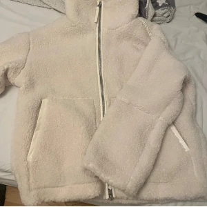 Beige teddyjacka med dragkedja - Mysig beige teddyjacka med dragkedja framtill och stora fickor. Perfekt för kyliga dagar när du vill hålla dig varm och stilren. Jackan har en hög krage och långa ärmar för extra komfort.