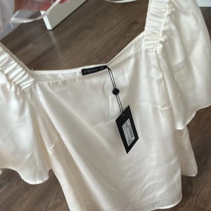 Vit blus från Stockh LM - Elegant vit blus från Stockh LM i ett glansigt material. Blusen har korta ärmar och en fyrkantig halsringning med rynkade detaljer vid axlarna. Perfekt för en stilren look.  Kunden står för frakten/ alternativt mötas upp i Norrköping!