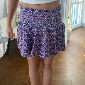 Blommig kortkjol i bomull - Säljer en söt blommig kortkjol i bomull med volangdetaljer. Kjolen har en härlig mix av blå, rosa och vit färg som ger en fräsch känsla. Perfekt för varma sommardagar!