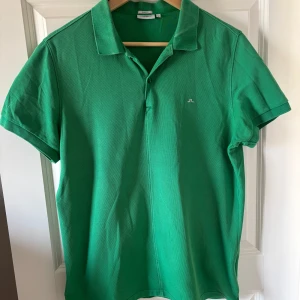 Grön pikétröja från J.Lindeberg - Säljer en snygg grön pikétröja från J.Lindeberg i storlek M. Tröjan har en klassisk krage och korta ärmar, perfekt för en stilren look. Den är tillverkad i ett bekvämt material och har en liten logga på bröstet. Passar bra till både jeans och shorts. Tröjan har en liten defekt på bröstet men inget som påverkar användningen. Hör av er vid frågor!