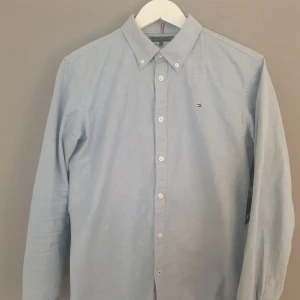 Ljusblå skjorta från Tommy Hilfiger - Säljer en stilren ljusblå skjorta från Tommy Hilfiger med klassisk krage och knappar framtill. Skjortan har långa ärmar och en liten logotyp på bröstet. Perfekt för en snygg och avslappnad look.