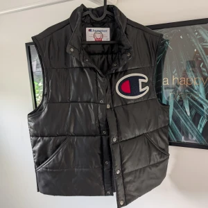 Svart dunväst från Supreme x Champion - Säljer en svart dunväst från Supreme i samarbete med Champion. Västen har en stor logga på bröstet och SUPREME tryckt på ryggen. Den är quiltad med knappar framtill och har två fickor.