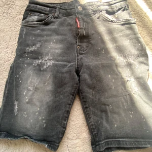 Svarta jeansshorts från Dsquared2 - Säljer ett par svarta jeansshorts från Dsquared2 med slitna detaljer och råa kanter. Shortsen har klassisk femficksdesign, knappgylf och en röd logotagg vid gylfen. Perfekta för en avslappnad stil.