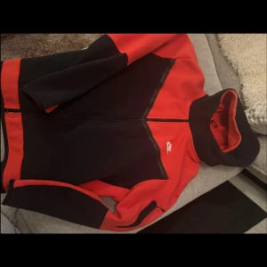 Nike svart och röd hoodie med dragkedja - Säljer en svart och röd hoodie från Nike med hel dragkedja och huva. Jackan har sportig design med kontrastpaneler och Nike-logga på bröstet. Perfekt för träning eller chill. Snygg och bekväm passform.