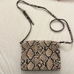 Axelväska i ormmönstrad syntet från H&M - Säljer en snygg axelväska från H&M i beige och brunt ormmönster. Väskan har lång justerbar axelrem och stängs med magnetknapp. Perfekt för att lyfta vilken outfit som helst med lite extra edge.