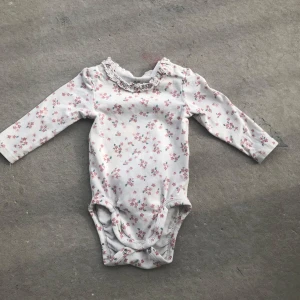 Vit blommig body med volang från H&M - Supersöt vit body från H&M med små rosa blommor och volangdetalj vid halsen. Långärmad modell med tryckknappar i grenen för enkel på- och avklädning. Perfekt för en gullig och bekväm look.