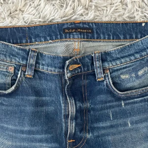 Blå jeans från Nudie Jeans Co - Säljer ett par klassiska blå jeans från Nudie Jeans Co med slitningar och orangea sömmar. Jeansen har fem fickor, normal passform och raka ben. Perfekta för dig som gillar en avslappnad stil med lite edge.