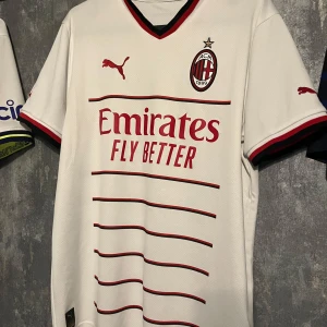 AC Milan vit bortatröja - Säljer en vit AC Milan bortatröja från Puma med röda och svarta detaljer. Tröjan har klubbmärke på bröstet, Puma-logga och Emirates Fly Better som sponsortryck. Tunn och luftig med korta ärmar och snygga ränder framtill.