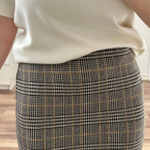 Rutig kjol storlek S - Säljer en kjol med rutigt mönster i svart, vitt och beige. Kjolen är figurnära och har en klassisk, stilren look. Midjehög modell. Använd ett fåtal gånger.