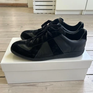 Svarta sneakers från Maison Margiela GAT - Säljer ett par helsvarta sneakers från Maison Margiela med både mocka- och skinnpaneler. Skorna har snörning och en platt sula, perfekt för en stilren och minimalistisk look. GATS